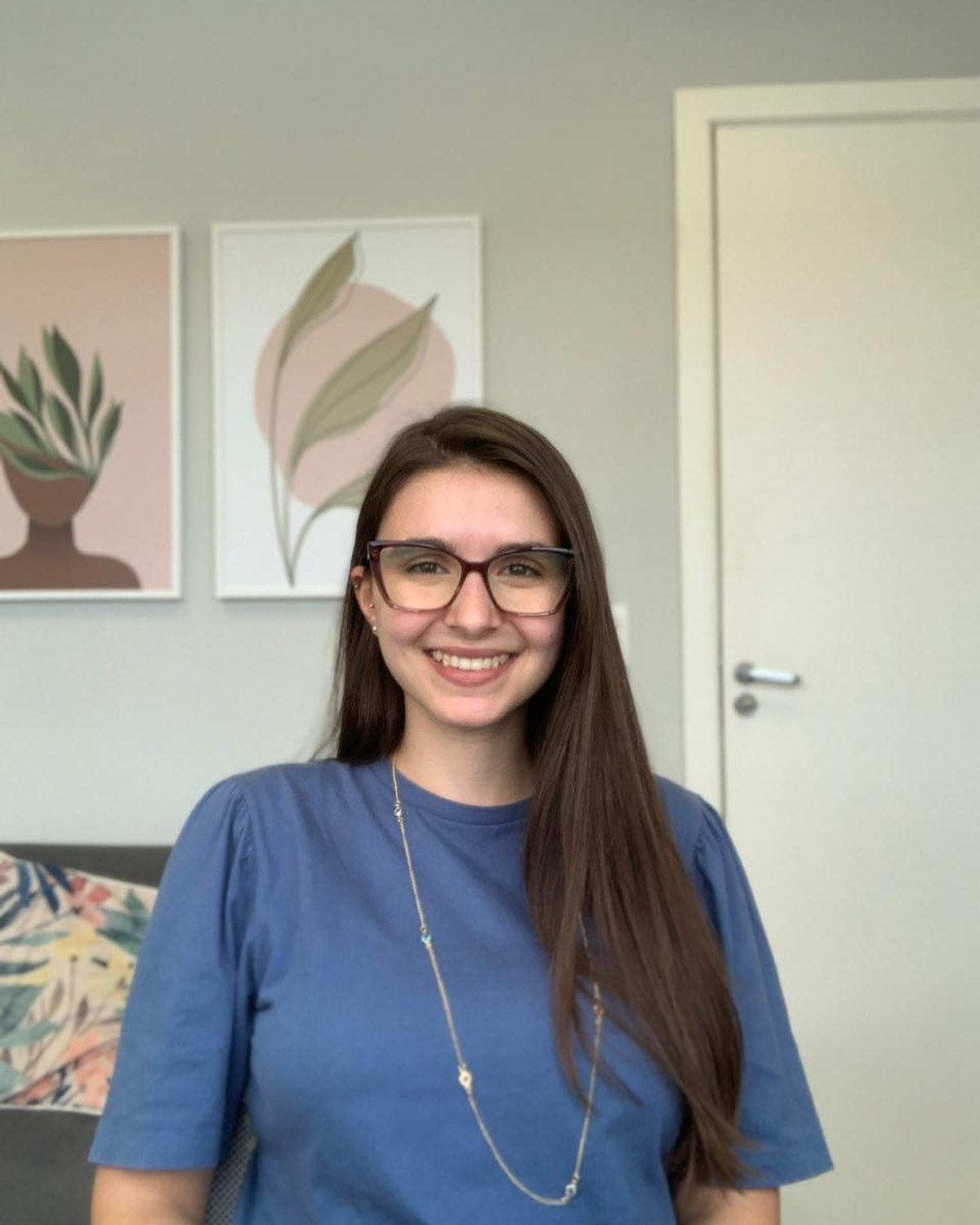 Mariana Saraiva, psicóloga, sorrindo de frente para a câmera, com camiseta azul e óculos, em ambiente acolhedor com artes botânicas no fundo.