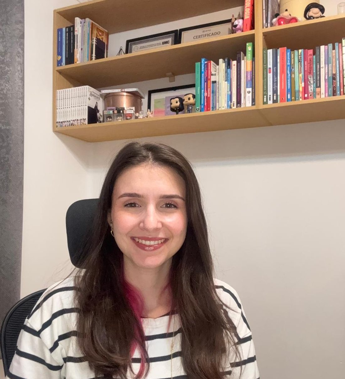 Mariana Saraiva sentada em sua sala de atendimento, sorrindo, em frente a uma estante de livros de psicologia e certificados acadêmicos.